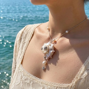 May include: A silver chain necklace featuring a large, iridescent seashell pendant adorned with small white pearls and intricate silver metal accents. The necklace includes additional pearls spaced along the chain, creating a layered, detailed look.