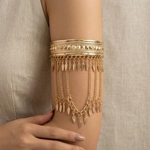 Op de afbeelding: Een goudkleurige metalen armmanchet met een brede, getextureerde band voorzien van een geometrisch piramidepatroon. Aan de onderrand hangen meerdere fijne kettinkjes, elk eindigend in een klein, plat, bladvormig bedeltje dat naar beneden valt.