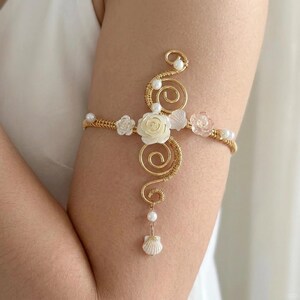 May include: A gold-toned wire arm cuff featuring a central cream-colored carved rose, smaller white and translucent pink floral accents, and several small white pearls. The design includes two prominent wire spirals and a dangling white shell charm.