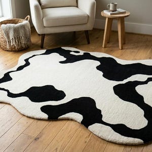 May include: An area rug featuring an irregular, organic shape with a high-contrast cow print pattern consisting of large, abstract black patches set against a solid cream-colored background, designed with a soft, textured pile surface.