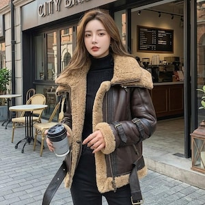 Könnte beinhalten: Braune Lederjacke mit beigem Shearling-Kragen und -Futter. Die Jacke hat einen Gürtel, Schnallen an den Ärmeln und einen Reißverschluss. Die Person trägt einen schwarzen Rollkragenpullover und hält eine Kaffeetasse.
