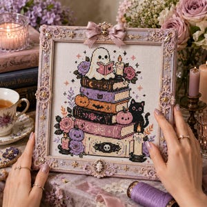 May include: A cross-stitch embroidery in a decorative lavender frame featuring a stack of books topped by a white ghost reading a book. The design includes a black cat, pumpkins, roses, and candles in shades of purple, orange, and black on fabric.