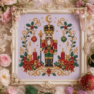 May include: A cross-stitch embroidery featuring a central nutcracker figure in a red, green, and gold uniform, flanked by red candles, holly branches, and ornaments on a light purple fabric, all encased in an ornate, cream-coloured frame.