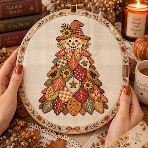 Pode incluir: Um bastidor de bordado circular contendo um design em ponto cruz de um espantalho com chapéu, cujo corpo é formado por um arranjo triangular de folhas de outono coloridas, girassóis e pequenos padrões sobre um tecido bege claro.