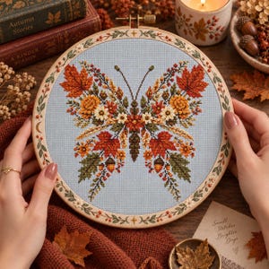 Op de afbeelding: Een ronde houten borduurring met een kruissteekontwerp van een vlinder op lichtblauwe stof. De vlindervleugels zijn samengesteld uit herfstbladeren, kleine bloemen en eikels in tinten oranje, rood, geel en groen.