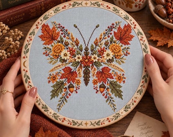 May include: A circular wooden embroidery hoop containing a cross-stitch design of a butterfly on light blue fabric. The butterfly wings are composed of autumn leaves, small flowers, and acorns in shades of orange, red, yellow, and green.