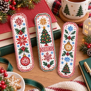 Puede incluir: Tres marcapáginas rectangulares de punto de cruz con motivos festivos. Los diseños incluyen un adorno rojo, un árbol de Navidad decorado y una bola dorada, rodeados de hojas de acebo verdes, bayas rojas y copos de nieve azules sobre tela blanca.