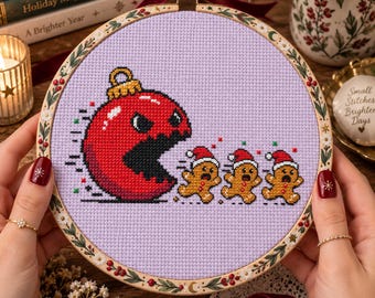 May include: A cross-stitch embroidery in a wooden hoop featuring a large red Christmas ornament with a wide open mouth chasing three small gingerbread cookies wearing red Santa hats, all stitched on a light purple fabric background.