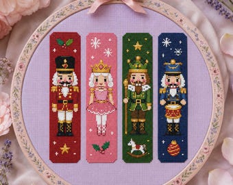 May include: A round wooden embroidery hoop containing a light purple fabric with four vertical cross-stitch bookmarks. The designs feature a red nutcracker, a pink ballerina, a green king, and a blue drummer, all accented with holiday motifs.