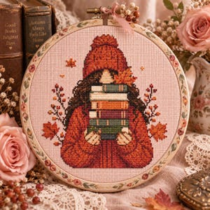 May include: A circular embroidery hoop containing a cross-stitch pattern of a person wearing a dark orange knit hat and sweater, holding a stack of five colorful books. The design features autumn leaves, branches, and a light pink fabric background.