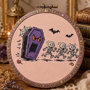 May include: A circular embroidery hoop containing a cross-stitch design on light pink fabric. The design features a purple coffin with a face chasing three white skeletons. Two black bats, a spider web, a small orange leaf, and candy are included.