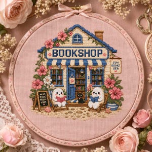 May include: A circular embroidery hoop containing a cross-stitch design of a bookshop on pink fabric. The shop features blue trim, a striped awning, and the text BOOKSHOP. Two white ghost-like figures read books, with a black cat and pink flowers.