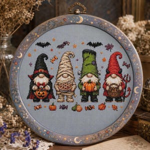 May include: A circular embroidery hoop containing a cross-stitch pattern of four Halloween gnomes on a light blue fabric. The gnomes are dressed as a vampire, a mummy, a monster, and a devil, surrounded by small pumpkins, bats, and candy icons.