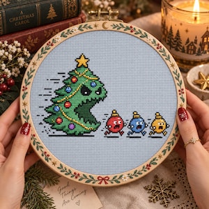 May include: A circular wooden embroidery hoop containing a cross-stitch design on light blue fabric. The design features a large green Christmas tree with a face chasing three small, colorful, frightened-looking Christmas ornament characters.