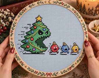 May include: A circular wooden embroidery hoop containing a cross-stitch design on light blue fabric. The design features a large green Christmas tree with a face chasing three small, colorful, frightened-looking Christmas ornament characters.