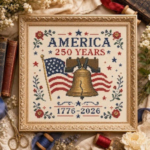 May include: A framed cross-stitch embroidery featuring the text AMERICA 250 YEARS and 1776-2026. The design includes a central Liberty Bell, an American flag, red flowers, and blue stars, all stitched in red, white, blue, and gold on cream fabric.