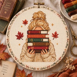 May include: A circular embroidery hoop containing a cross-stitch pattern of a person with a messy bun holding a stack of five books. The design features red maple leaves, small yellow stars, and a cream-colored sweater. The hoop frame has a floral border.