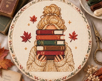 May include: A circular embroidery hoop containing a cross-stitch pattern of a person with a messy bun holding a stack of five books. The design features red maple leaves, small yellow stars, and a cream-colored sweater. The hoop frame has a floral border.