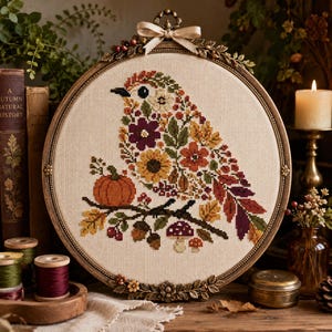 May include: A circular cross-stitch embroidery featuring a bird composed of autumn-colored flowers, leaves, and berries. The bird sits on a branch with a small orange pumpkin and mushrooms. The piece is framed in an ornate bronze-toned hoop.