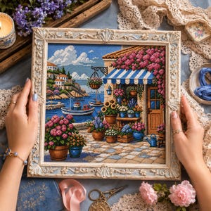 May include: A framed cross-stitch embroidery depicting a Mediterranean coastal scene with a flower shop, a blue and white striped awning, potted plants, and a boat on the water. The ornate, light-colored frame features decorative floral carvings.