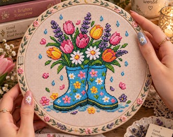 Op de afbeelding: Een ronde borduurring met een kruissteekontwerp van blauwe regenlaarzen gevuld met een boeket van roze, gele en oranje tulpen, witte madeliefjes en paarse lavendeltakjes, versierd met kleine geborduurde bloemetjes op de rand.