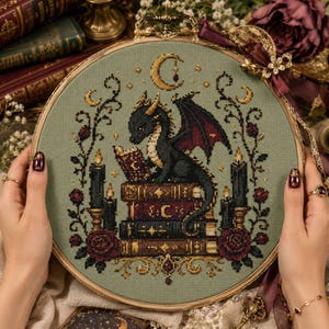 May include: A circular wooden embroidery hoop containing a cross-stitch design on sage green fabric. The pattern features a black dragon reading a book while perched on a stack of three books, flanked by black candles, red roses, and a crescent moon.