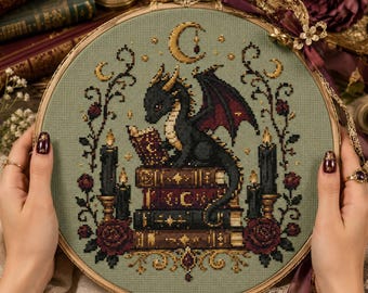 May include: A circular wooden embroidery hoop containing a cross-stitch design on sage green fabric. The pattern features a black dragon reading a book while perched on a stack of three books, flanked by black candles, red roses, and a crescent moon.