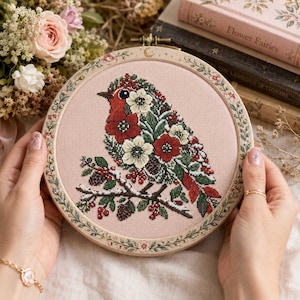 May include: A circular embroidery hoop containing a cross-stitch design of a bird composed of red and white flowers and green leaves. The bird sits on a branch with pinecones and red berries against a light pink fabric background.