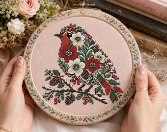 May include: A circular embroidery hoop containing a cross-stitch design of a bird composed of red and white flowers and green leaves. The bird sits on a branch with pinecones and red berries against a light pink fabric background.