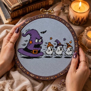 May include: A circular embroidery hoop containing a cross-stitch design on light blue fabric. The design features a large, angry purple witch hat chasing three small, frightened white ghosts wearing colorful hats, with a small black bat above.