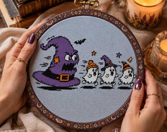 May include: A circular embroidery hoop containing a cross-stitch design on light blue fabric. The design features a large, angry purple witch hat chasing three small, frightened white ghosts wearing colorful hats, with a small black bat above.