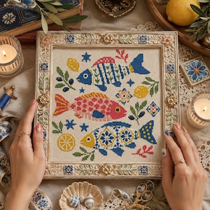May include: A square cross-stitch embroidery featuring three stylized fish in blue, pink, and yellow, surrounded by lemon and olive branch motifs, encased in an ornate, distressed cream-colored wooden frame with floral and geometric accents.