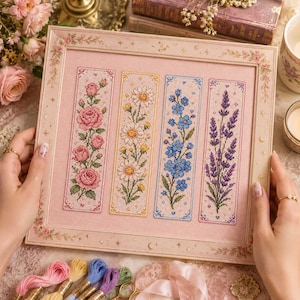 Könnte beinhalten: Eine rechteckige gerahmte Kreuzstichstickerei mit vier vertikalen Paneelen auf hellrosa Gitterstoff. Die Paneele zeigen florale Motive: rosa Rosen, weiße Gänseblümchen, blaue Vergissmeinnicht und lila Lavendel, eingefasst in einen dekorativen Rahmen.