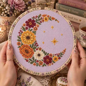 May include: A circular wooden embroidery hoop containing a cross-stitch design on light purple fabric. The pattern features a crescent moon shape formed by various flowers in shades of yellow, orange, red, and white, accented with small yellow stars.