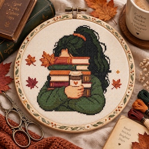 May include: A circular embroidery hoop containing a cross-stitch design of a person with dark green hair holding a stack of books and a coffee cup. The design features autumn leaves and is framed by a wooden hoop decorated with painted floral vines.