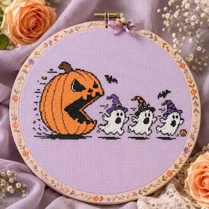 Puede incluir: Un bastidor de bordado circular con un diseño de punto de cruz sobre tela morada clara. El diseño muestra una gran calabaza naranja persiguiendo a tres pequeños fantasmas blancos con sombreros de bruja, junto a pequeños murciélagos negros.