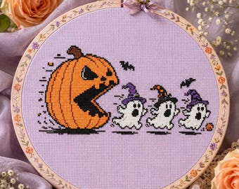 May include: A circular embroidery hoop containing a cross-stitch design on light purple fabric. The design features a large orange jack-o'-lantern chasing three small white ghosts wearing purple and orange witch hats, with small black bats.