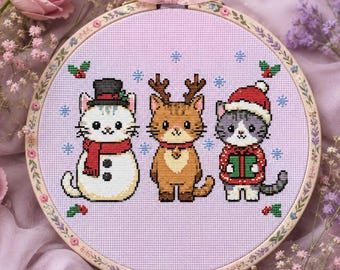 May include: A circular embroidery hoop containing a cross-stitch pattern on light purple fabric. The design features three cats: one dressed as a snowman with a top hat and scarf, one with reindeer antlers, and one wearing a Santa hat holding a gift.