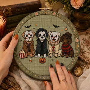 May include: A circular embroidery hoop containing a cross-stitch design on sage green fabric. The design features four dogs dressed as horror movie characters: Jason Voorhees, a dark-haired figure, Pennywise, and Freddy Krueger, with a pumpkin.