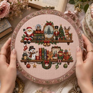 Op de afbeelding: Een ronde borduurring met een kruissteekontwerp op lichtroze stof. Het patroon toont twee houten planken gevuld met kerstmotieven, waaronder de kerstman, een sneeuwpop, een notenkraker, een rendier, boeken en cadeautjes.