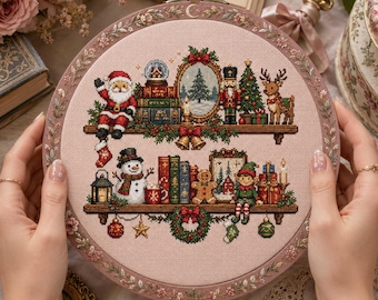 Pode incluir: Um bastidor de bordado circular contendo um design em ponto cruz sobre tecido rosa claro. O padrão apresenta duas prateleiras de madeira repletas de motivos natalinos, incluindo o Papai Noel, um boneco de neve, um quebra-nozes, uma rena, livros e presentes.