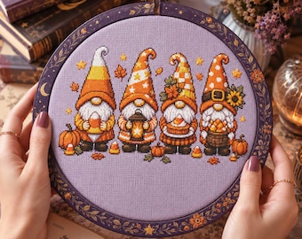May include: A circular embroidery hoop containing a cross-stitch pattern of four gnomes on a light purple fabric. The gnomes wear orange and yellow hats with autumn-themed motifs, including pumpkins, sunflowers, lanterns, and candy corn accents.