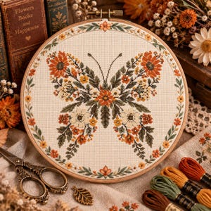 Op de afbeelding: Een ronde houten borduurring met een kruissteekontwerp van een vlinder gevormd door oranje, gele en witte bloemen met groene bladeren. Het ontwerp is omlijst door een bloemenrand op crèmekleurige stof met een metalen schroefsluiting.