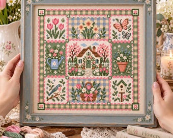 May include: A square cross-stitch embroidery in a blue wooden frame featuring a central cottage surrounded by garden scenes including tulips, daffodils, butterflies, a blue bird, a watering can, a flower basket, and a birdhouse on a blue chequered background.