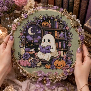 Peut inclure: Un tambour à broder circulaire présentant un point de croix représentant un fantôme blanc lisant un livre violet dans une bibliothèque. La scène inclut un chat noir, des citrouilles, des bougies et une fenêtre avec un croissant de lune.