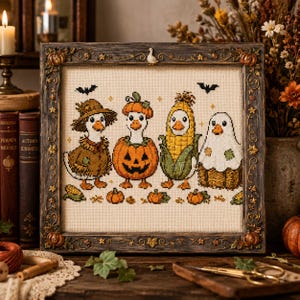 Könnte beinhalten: Ein gerahmtes Kreuzstichbild auf cremefarbenem Stoff mit vier Enten in Halloween-Kostümen: eine mit braunem Hut, eine in einem Kürbis, eine als Maiskolben verkleidet und eine als Geist. Über den Enten sind zwei schwarze Fledermäuse eingestickt.