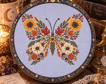Può includere: Un telaio da ricamo circolare contenente un disegno a punto croce a forma di farfalla composto da girasoli, foglie autunnali, fiori bianchi e ghiande su tessuto azzurro. La cornice in legno scuro presenta motivi dorati di lune e stelle.
