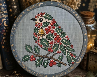 May include: A circular embroidery hoop containing a cross-stitch pattern of a bird perched on a holly branch. The bird is decorated with red berries and green leaves, featuring a small gold bell. The frame is dark blue with gold stars and moons.
