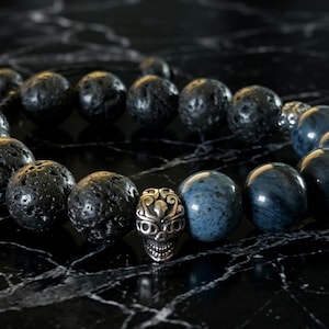 May include: A beaded bracelet featuring dark gray lava rock beads, blue-gray stone beads, and a silver skull charm. The bracelet is displayed on a black marble surface, creating a striking contrast. The bracelet is a unique accessory.