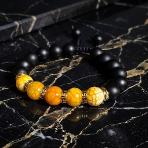 May include: A beaded bracelet featuring black onyx and yellow-orange gemstone beads. The bracelet has gold-colored accents and is displayed on a black marble surface. The bracelet is adjustable.